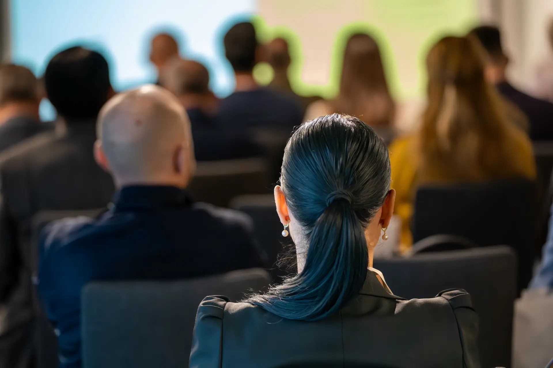 A group of people attending a conference, viewed from the back, focusing on a woman with a ponytail